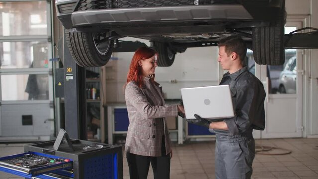 car maintain, professional young mechanic in uniform tells pleased female owner of vehicle about condition of running machine on hydraulic ramp at technical platform