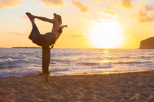 Silhouette of young woman practicing standing bow yoga pose on sandy beach at sunset.