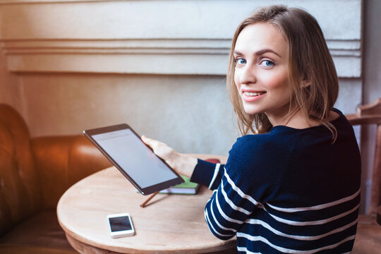 Young female student is enjoying free time,while is sitting with touch pad in coffee shop indoor. Beautiful woman is holding digital tablet and looking at camera during rest in cafe.