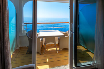 A cruise ship stateroom balcony with a table and two large armchairs overlooking a blue Mediterranean Sea seen through an open sliding glass door.