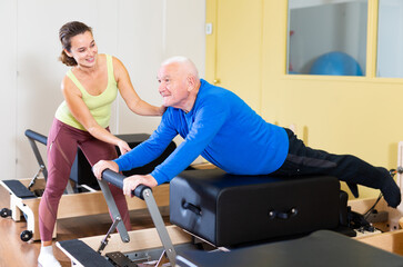 Female instructor of pilates helping elderly man workout. Two people working in pilates studio, woman assistant supporting and correcting old man patient beginner.