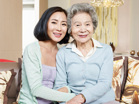 asian mother and adult daughter sitting on couch smiling