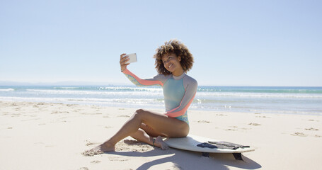 Smiling female taking selfie on smartphone sitting on surfboard on beach. Tarifa beach. Provincia Cadiz. Spain.