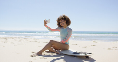 Smiling female taking selfie on smartphone sitting on surfboard on beach. Tarifa beach. Provincia...