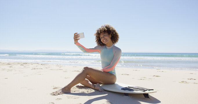 Smiling female taking selfie on smartphone sitting on surfboard on beach. Tarifa beach. Provincia Cadiz. Spain.