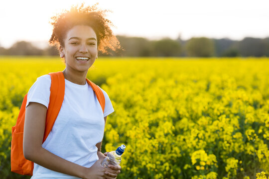Outdoor portrait of beautiful happy mixed race African American girl teenager female young woman with water bottle hiking with back pack in a field of yellow flowers at sunset in golden evening sunshi