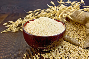 Oat flour in a bowl, bag with oat and stalks of oats on the background of dark wood planks