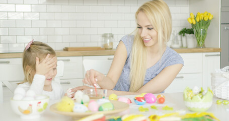 Fototapeta na wymiar Laughing young mother and little adorable girl coloring Easter eggs together while sitting at table in kitchen.