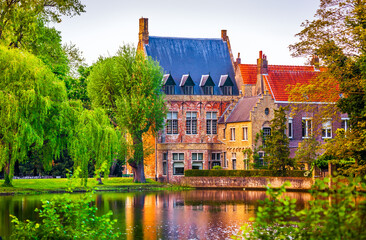 Fototapeta premium Vintage building over lake of love in Minnewater park in Bruges Belgium near Beguinage monastery of Beguines. Picturesque landscape with green trees sunset time.