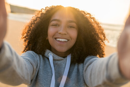 Outdoor portrait of beautiful happy mixed race African American girl teenager female young woman on a beach taking a selfie photograph smiling laughing with perfect teeth in golden sunset evening suns