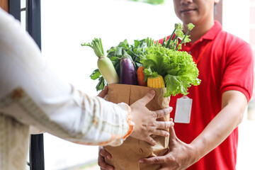 Asian deliver man in red uniform handling bag of food, fruit, vegetable give to female costumer in...