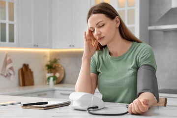 Woman suffering from headache and measuring blood pressure in kitchen, space for text