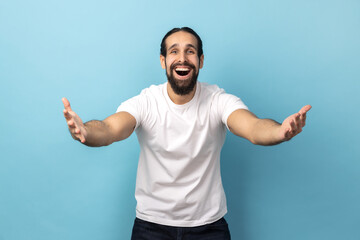 Naklejka premium Welcome, wide open hug. Portrait of man with beard wearing white T-shirt stretching hands to camera and smiling broadly, going to embrace, share love. Indoor studio shot isolated on blue background.