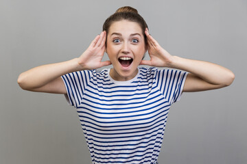 Portrait of excited amazed woman wearing striped T-shirt standing with hands on head, looking at camera with big eye, saying wow. Indoor studio shot isolated on gray background.