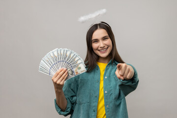 Happy angelic young woman with halo above head holding dollar banknotes and pointing to camera,...