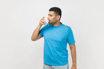 Portrait of thirsty handsome unshaven man in blue T- shirt standing with glass in hands, drinking...