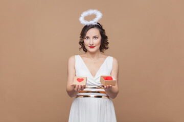 Portrait of beautiful middle aged woman with wavy hair and nimb over head, holding little present box, looking at camera, wearing white dress. Indoor studio shot isolated on light brown background.