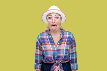 Portrait of childish mature woman wearing checkered shirt, hat and eyeglasses sticking out tongue,...