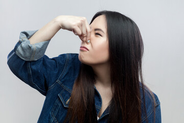 Portrait of disgusted brunette woman in blue denim jacket standing plugs nose as smells something stink and unpleasant, feels aversion. Indoor studio shot isolated on gray background. - obrazy, fototapety, plakaty