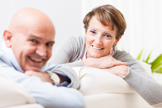 Relaxed friendly middle-aged couple leaning over the back of a comfortable sofa smiling at the camera with focus to the woman