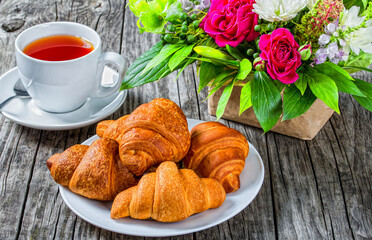 French crescent-shaped rolls - croissants on white dish, cup of tea and bouquet of fresh flowers and a gift wrapped  with paper on old wooden boards, close-up