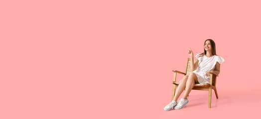 Pretty young woman in wooden armchair pointing at something on pink background with space for text