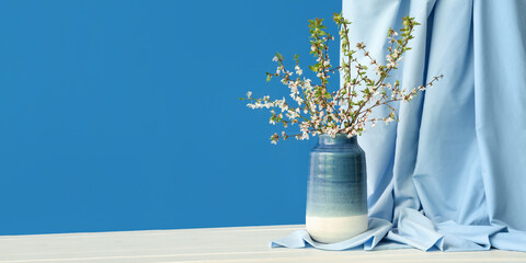 Vase with blooming tree branches and curtain on white table against blue background. Banner for...