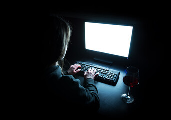 Silhouette of woman typing at computer in dark illuminated by screen with wine glass