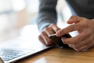 Close-up of incognito male hands holding black cell phone and scrolling the screen while sitting next to laptop on wooden table. Copy space