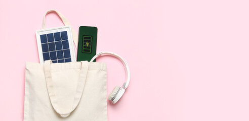 Bag with portable solar panel, mobile phone and headphones on pink background with space for text