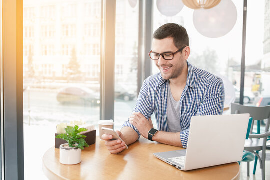 Coffee time. Handsome young man in cafe with big window. Man with cup of coffee to go. Man with glasses and laptop using mobile phone and smiling