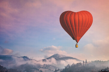 Beautiful red air balloon heart shape against blue and pink pastel sky in a sunny bright morning. Foggy mountains in the background. Romantic trip on Valentine's Day. Sport and recreation travel theme
