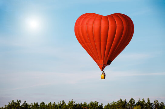 Beautiful red air balloon in the shape of heart against blue sky in a sunny bright day fly high above the trees. Romantic trip on Valentine's Day. Sports and recreation travel theme. Nature background