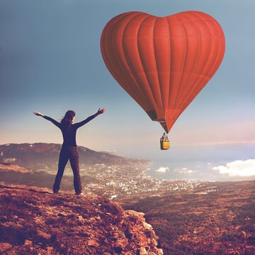 Girl with rised hands on the top of mountain looking at big air balloon in the sky. Romantic postcard background on Valentine's Day. Sport and recreation travel theme. Nature background. Back view