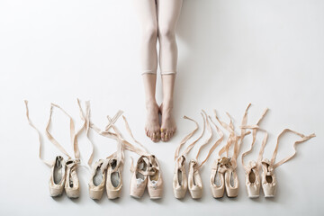 Many pairs of ballet shoes in pairs stand in a row. Legs of a young ballerina with bare feet. The girl's fingers are all covered in wounds and plasters. Pointe shoes in different condition from new to