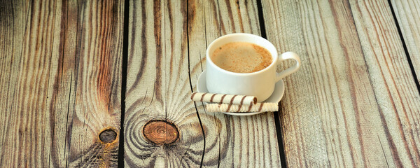 A cup of fresh cappuccino on a saucer and two waffle rolls on a light wooden table.