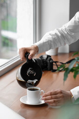 Fototapeta na wymiar Woman pouring coffee from pot into cup on wooden windowsill