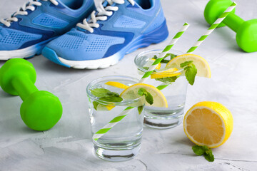 Sport composition with blue sneakers, green dumbbells, glasses of water with lemon, mint and straws...
