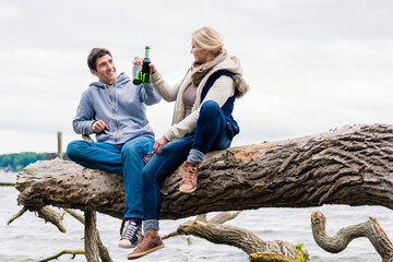 Young couple, woman and man, sitting on tree stump at the riverside drinking beer and saying cheers
