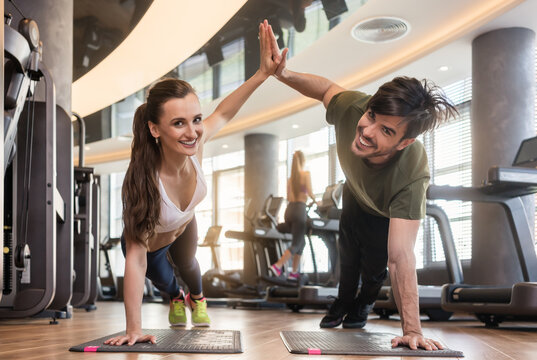 Young fit man and woman smiling and looking at camera while giving high five from basic plank pose during workout at the gym