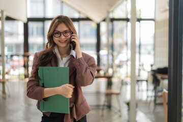 Fototapeta premium Businesswoman standing and holding a folder while talking smartphone in an office.