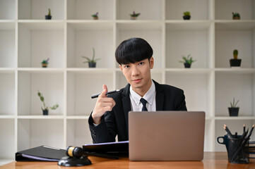 A smart Asian male lawyer in a formal suit points his finger at the camera while sitting at his desk