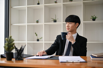 A handsome Asian male lawyer in a formal suit is sipping his morning coffee at his desk.