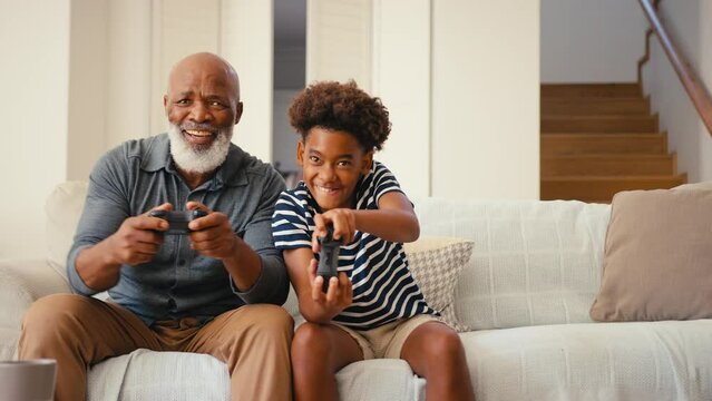 Grandfather and grandson sitting on sofa at home holding controllers playing video game together giving each other high five with multi-generation family in background - shot in slow motion