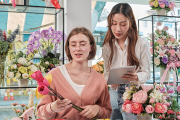 Two young beautiful female florist partners discuss bloom bouquet arrangement design in tablet, online purchase order work in colorful flower shop store with fresh flora, SME business entrepreneur. - obrazy, fototapety, plakaty