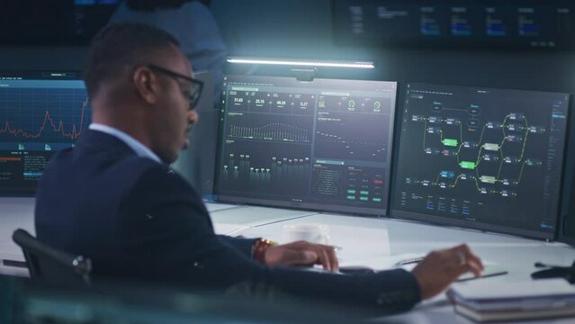 African American IT technical support specialist sits in front of computer with data server and real-time analysis charts in modern monitoring office, writes notes. Big digital screens on background.