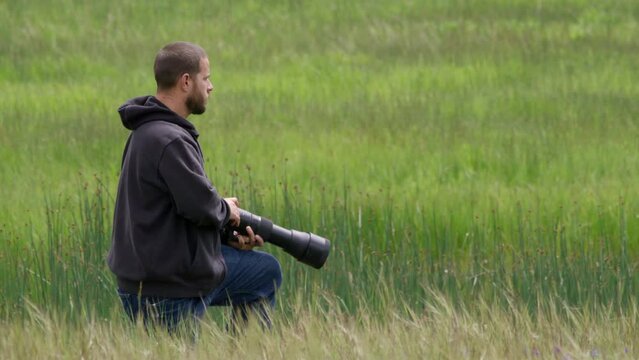Young adventure photographer taking photographs in wildlife environment. Male photographer, preparing to shoot some wildlife animals. Hiding in the bushes with big telephoto lens. 4K slow. motion