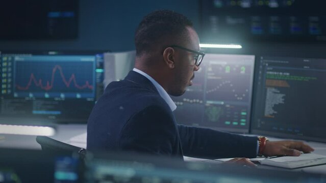 African American software engineer sits in front of computer and big digital screens with data server and blockchain network, works in modern monitoring control room with real-time analysis charts.