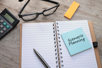 The word Scenario Planning is written on the Post-it Note on the empty notebook with pen, calculator, and eyeglasses on the wooden table