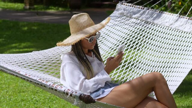 Woman traveler in straw hat and sunglasses lies on hammock and summer resort and hold mobile phone in hand. Woman traveler enjoying resting on hammock and using mobile phone app while summer travel